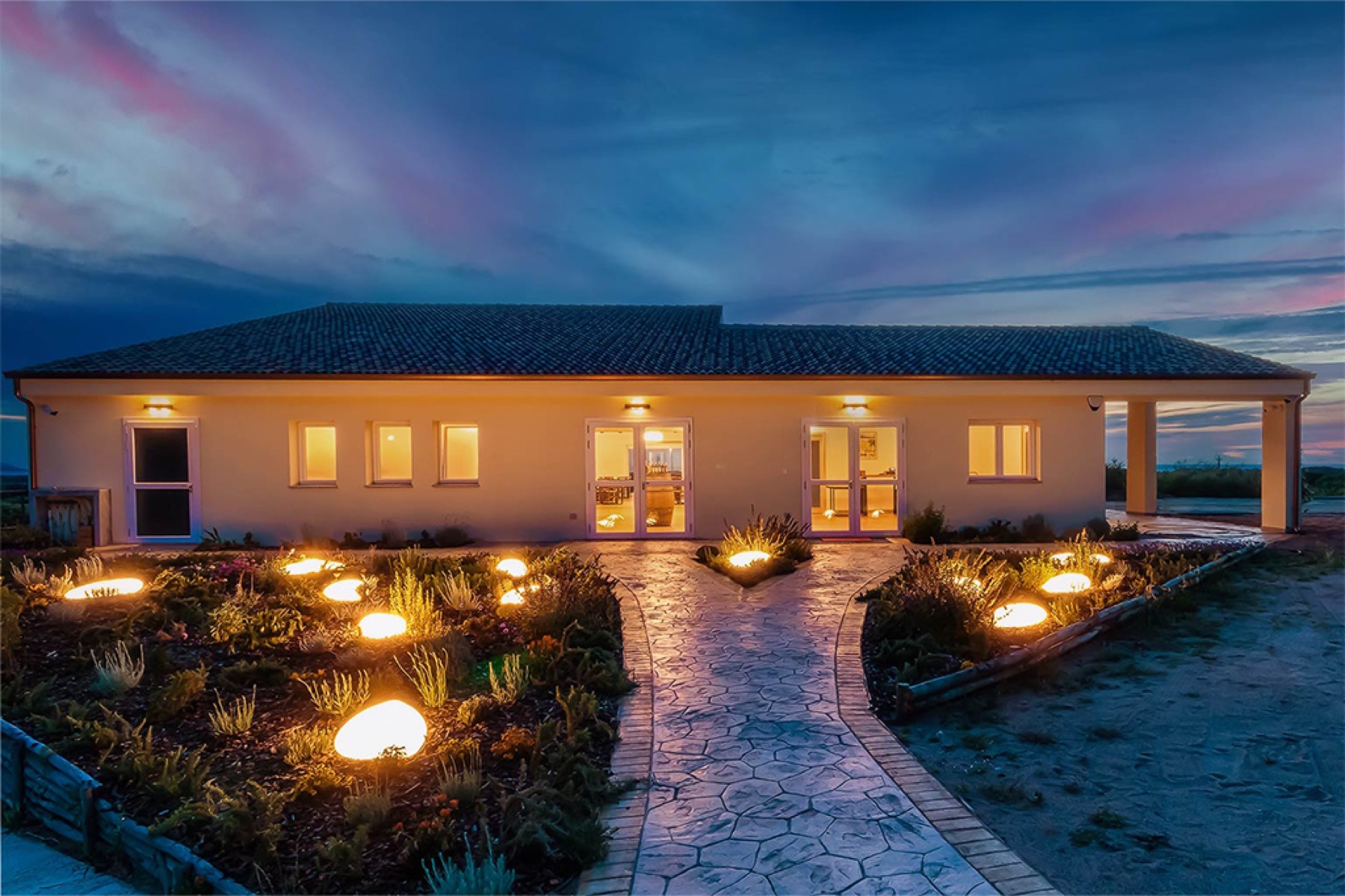 Cantina Li Duni's winery building at dusk, garden lights leading down to the entrance